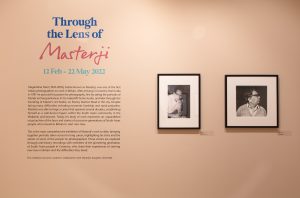 Photo of exhibition wall, Through the lens of Masterji. Two frames beside the text. Main title in blue, and Masterji in pink.